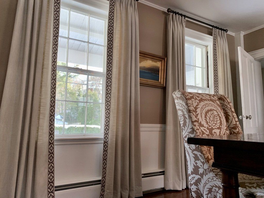 Living room with beige curtains, two windows, framed art, and a patterned chair beside a dark table