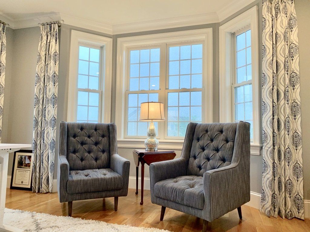 Two gray tufted armchairs in front of a bright bay window with patterned curtains.