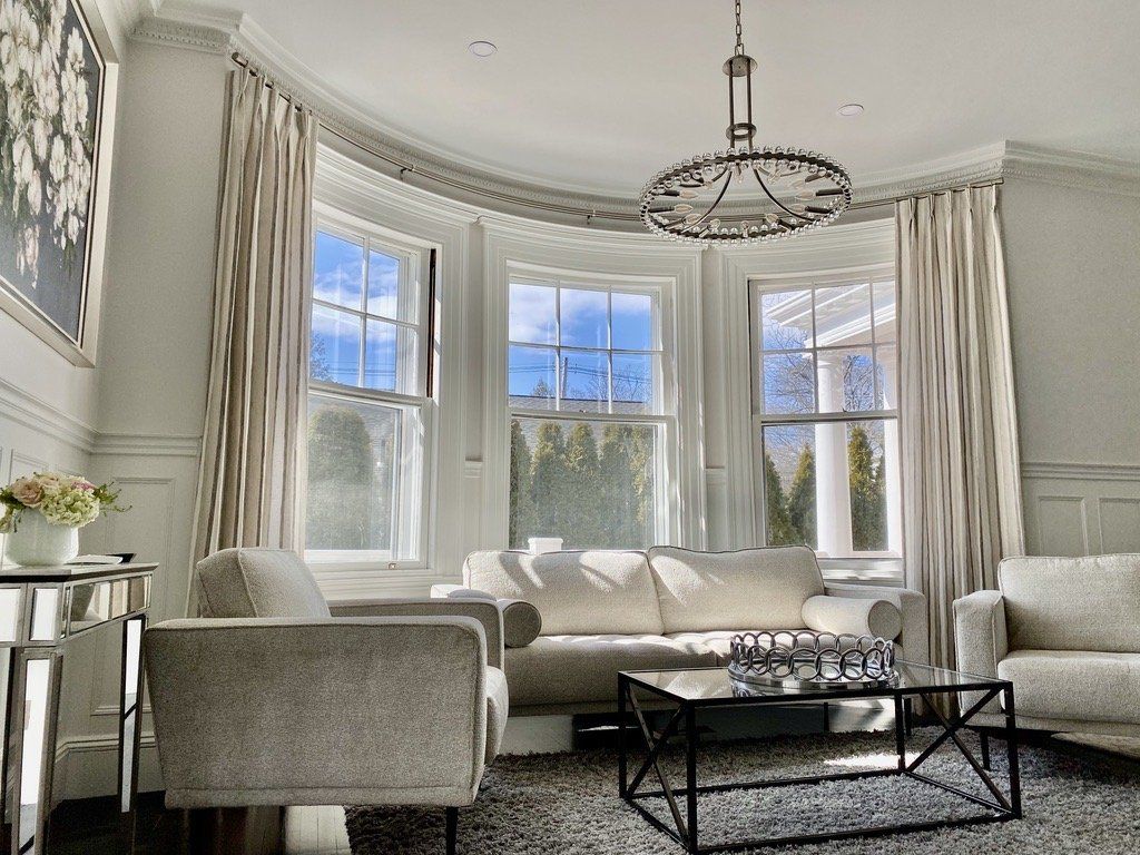 Elegant living room with bay windows, neutral-toned furniture, ornate crown molding, and a chandelier.