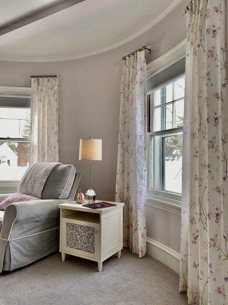 Cozy living room with gray couch, white nightstand, floral curtains, and a lamp.