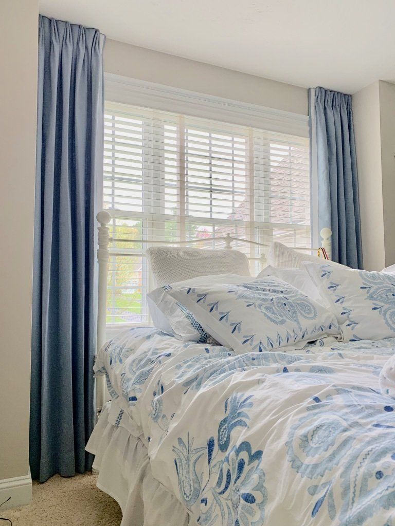 Bedroom with blue curtains, white blinds, and a bed with blue and white bedding.