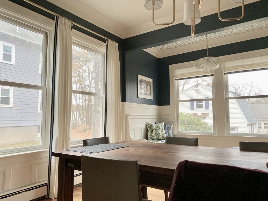 Dining room with a long wooden table, chairs, and windows with white curtains.