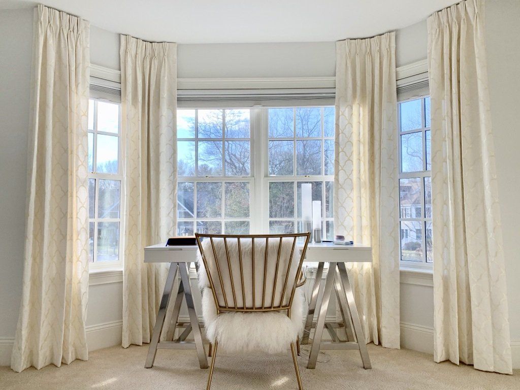 A bright home office with a white desk, gold chair, and three windows framed by cream curtains.