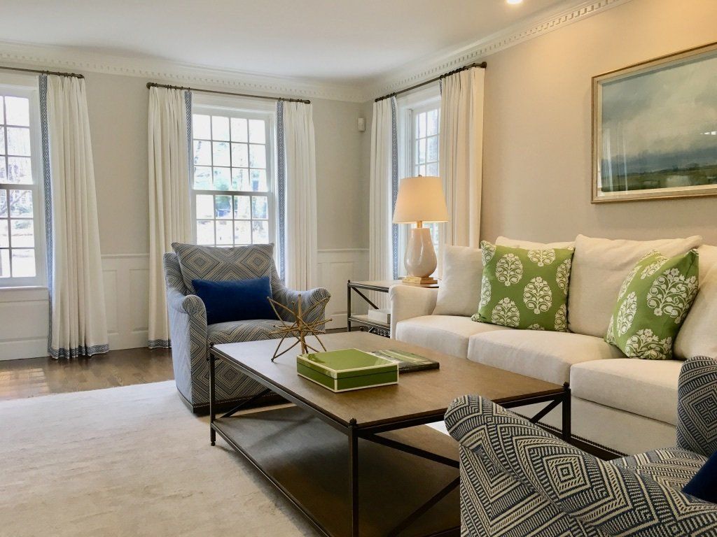 Living room with white sofa, patterned armchairs, and beige walls, lit by natural light.