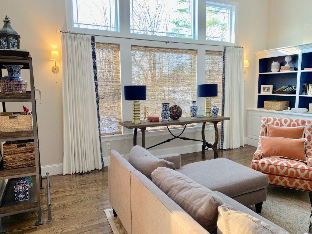 Living room with a sofa, ottoman, and console table in front of windows, flanked by bookshelves and a patterned chair.