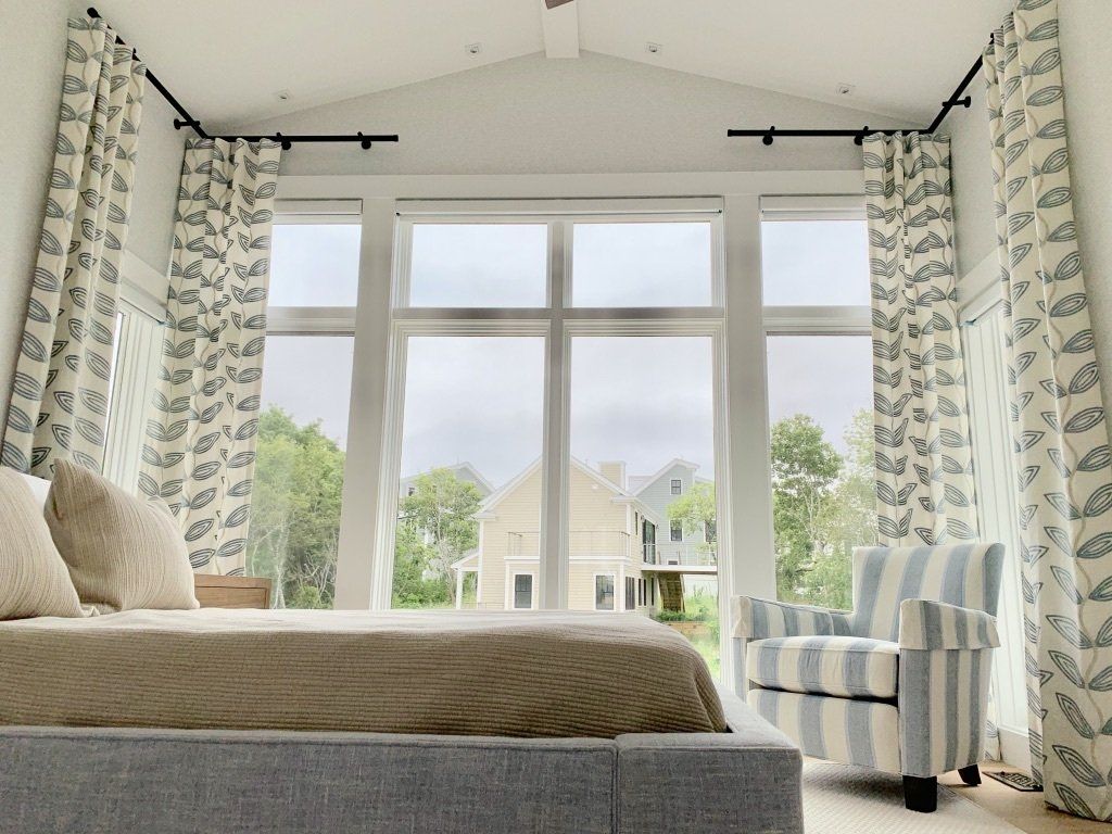 Bedroom with large window overlooking a house, bed, and striped armchair; patterned curtains.