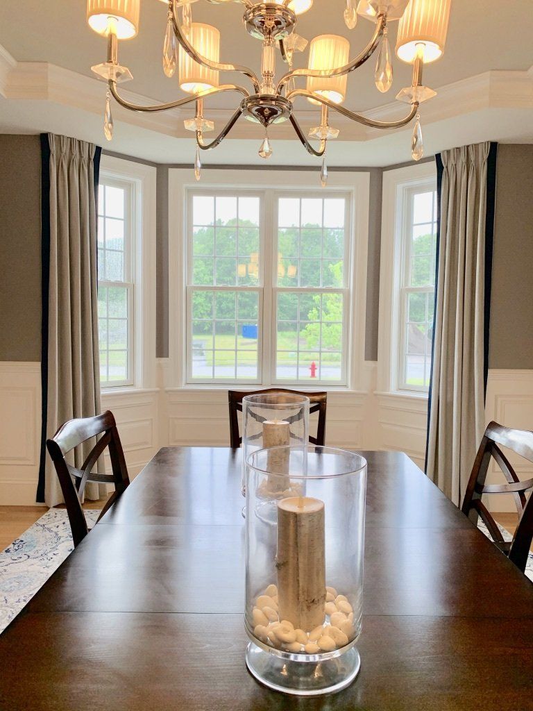 Dining room with a long wooden table, bay window, chandelier, and gray curtains.