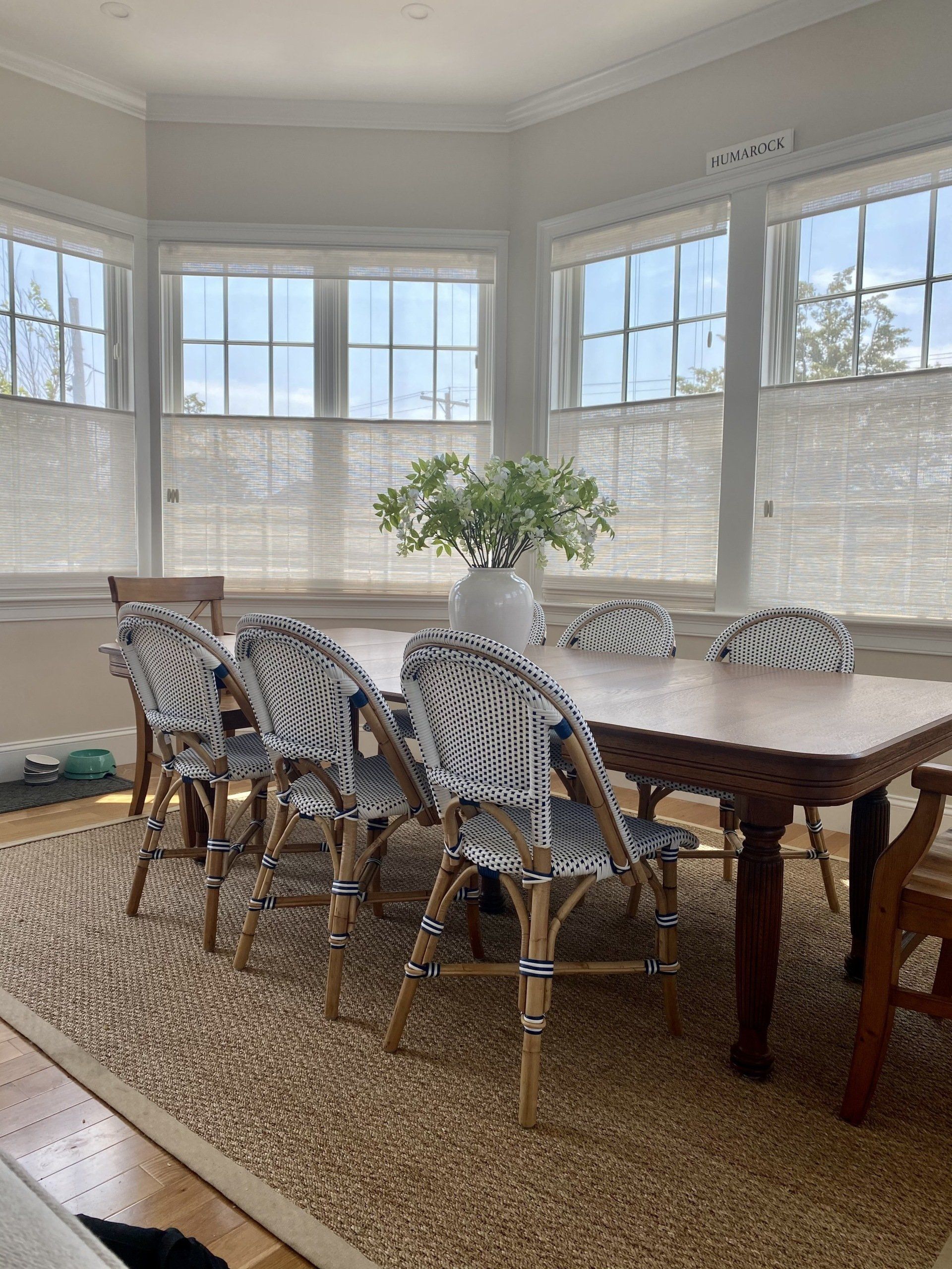 Dining room with wooden table, blue patterned chairs, and large vase of greenery in front of windows with blinds.