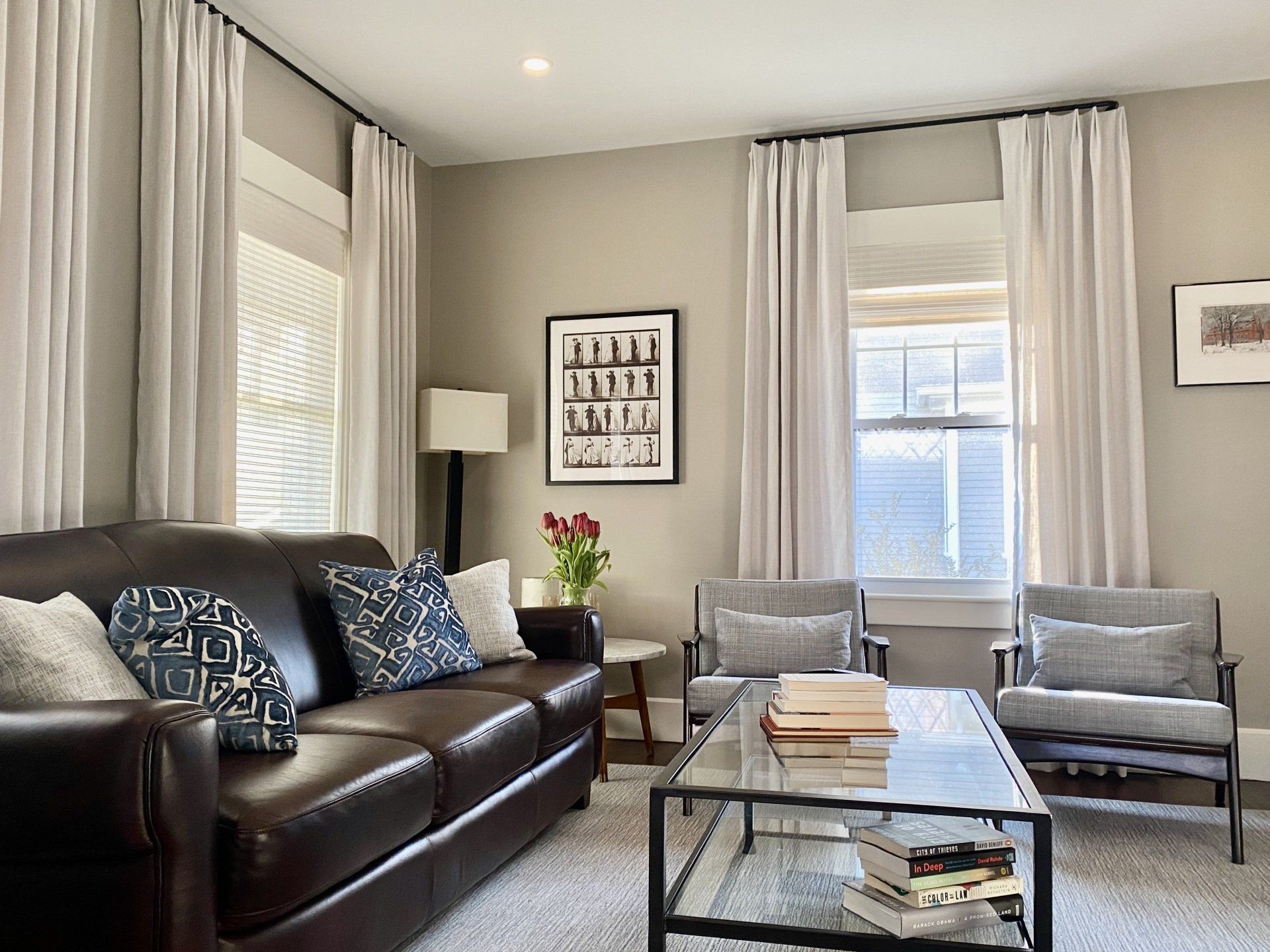 Living room with brown leather sofa, two gray armchairs, white curtains, and artwork.