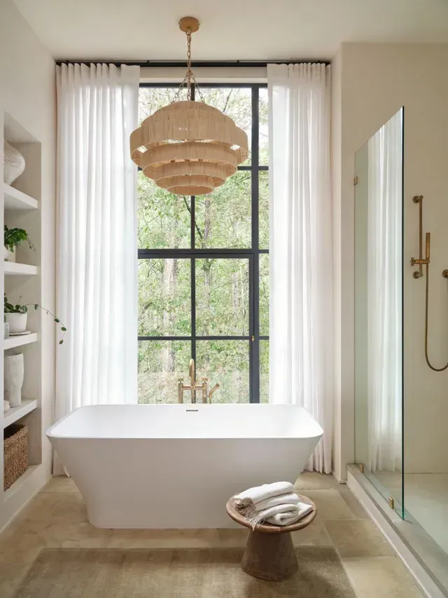 Bathroom with a white tub, large window, and woven chandelier. Sheer curtains frame the window with a lush forest view.