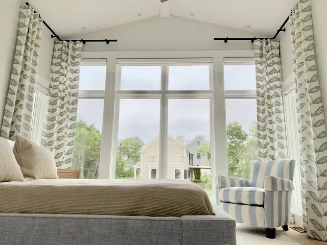 Bedroom with large windows, bed, striped armchair, and patterned curtains. Exterior view of trees and building.