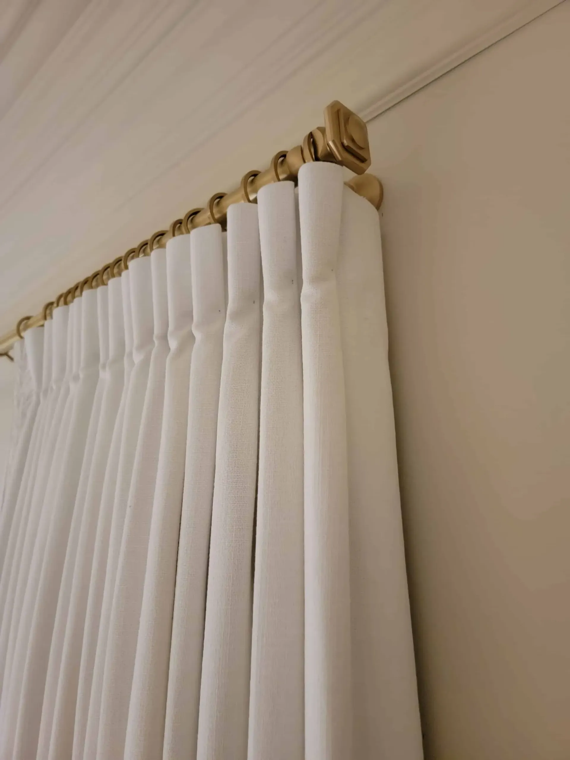 White vertical curtains hanging from a gold curtain rod against a beige wall and ceiling corner