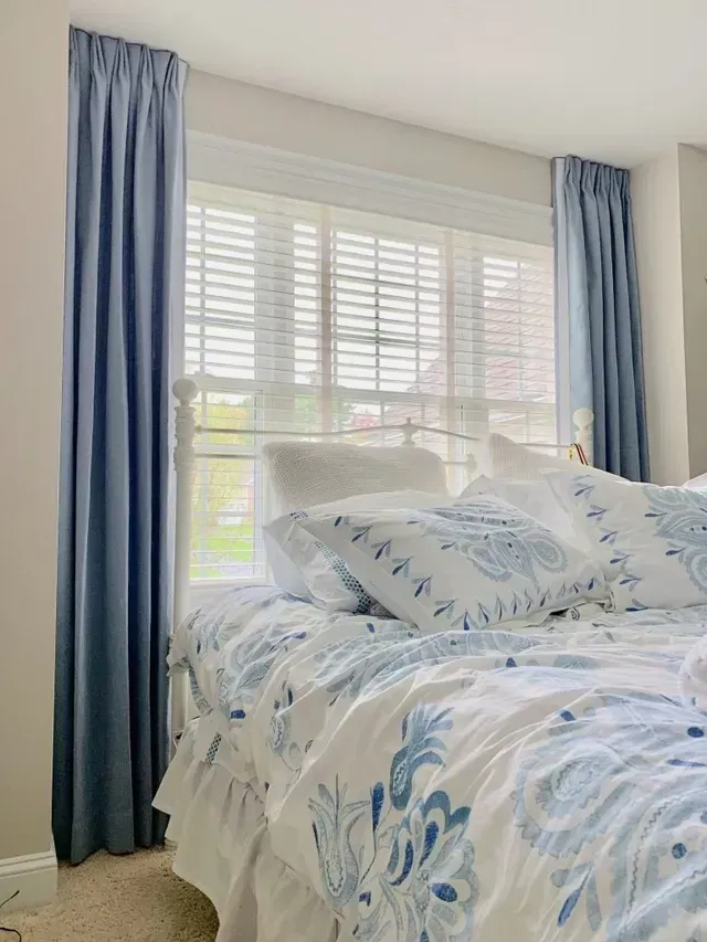 Bedroom with blue curtains, white blinds, and a bed with blue and white patterned bedding.