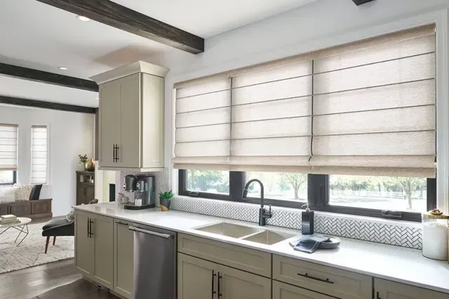 Kitchen with Roman shades, light-colored cabinets, countertop, sink, and window overlooking a green area.
