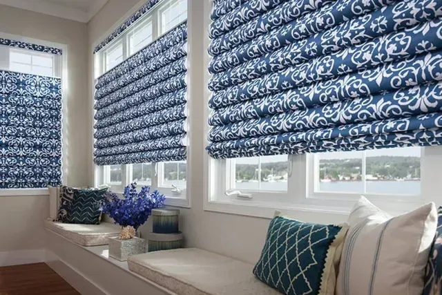 Blue patterned Roman shades in a sunroom with white window frames and window seat with pillows.
