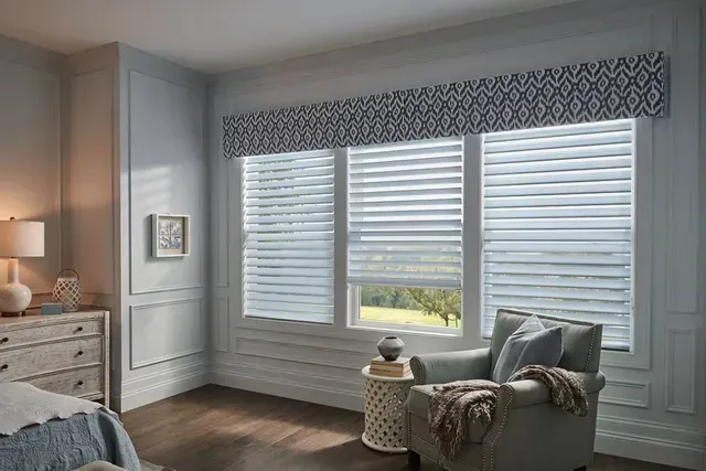 Bedroom with window shades, patterned valance, chair, side table, and dresser.