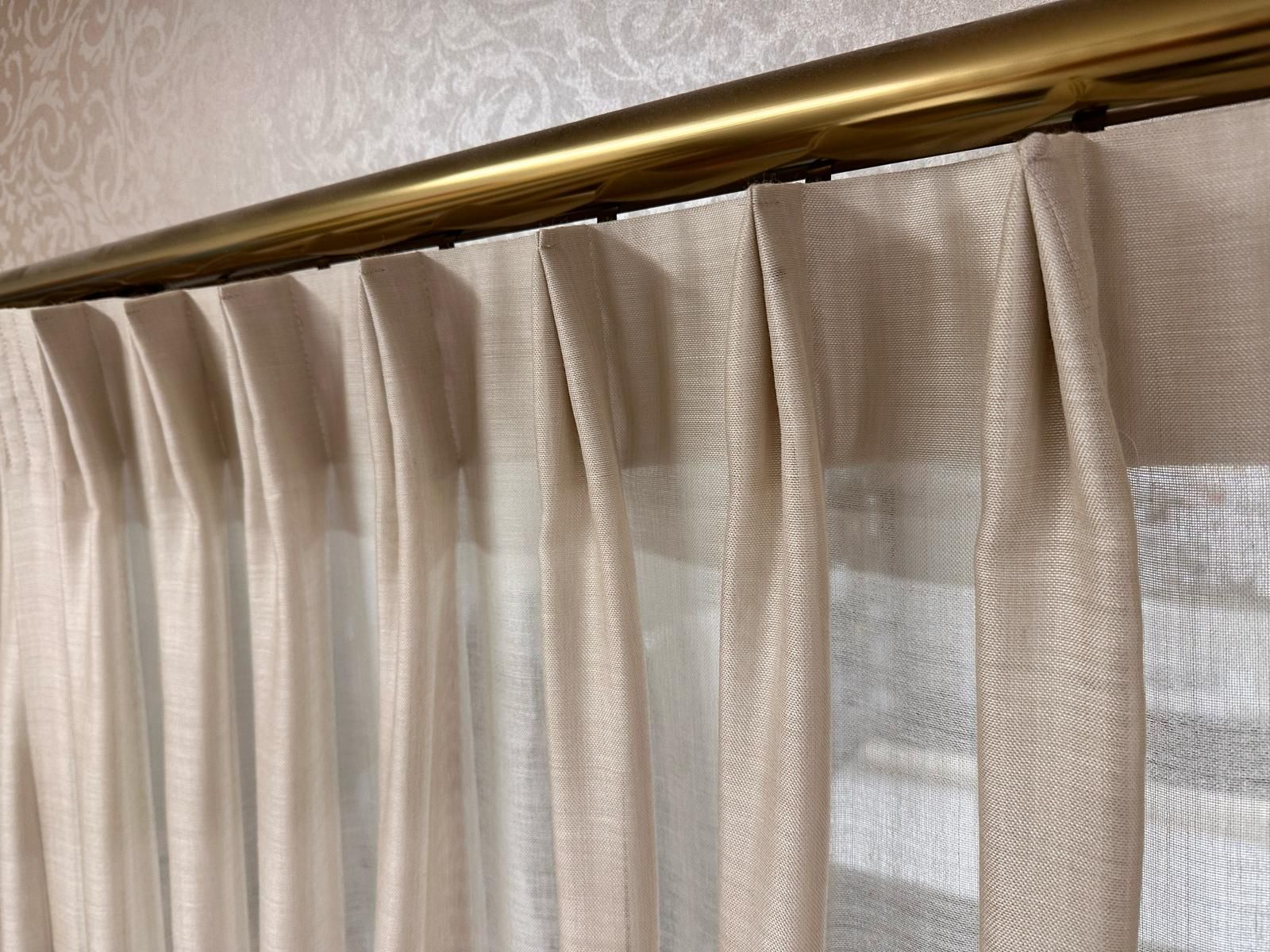 Beige pleated curtains hanging from a brass curtain rod by a window