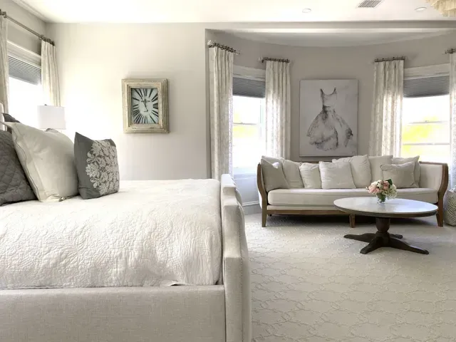 Bedroom with white bed, couch, and circular coffee table on light carpet. Gray walls, horse art.