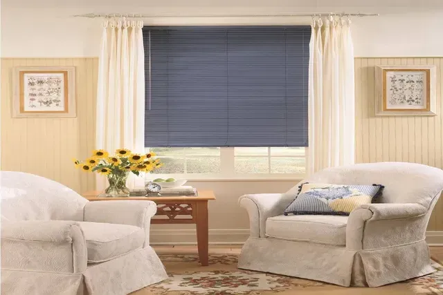 Cozy living room with blue blinds, white curtains, armchairs, small table, and framed art.