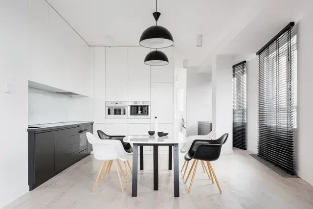 Modern white kitchen and dining area with black accents; includes cabinetry, table, chairs, and light fixtures.