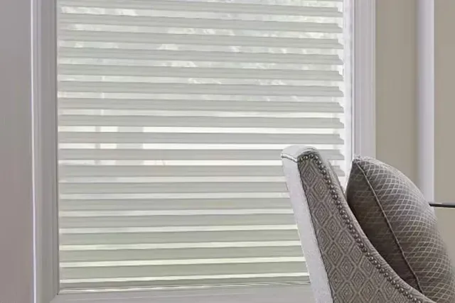 White layered window blinds, chair in front of a window. Light from outside filters through.