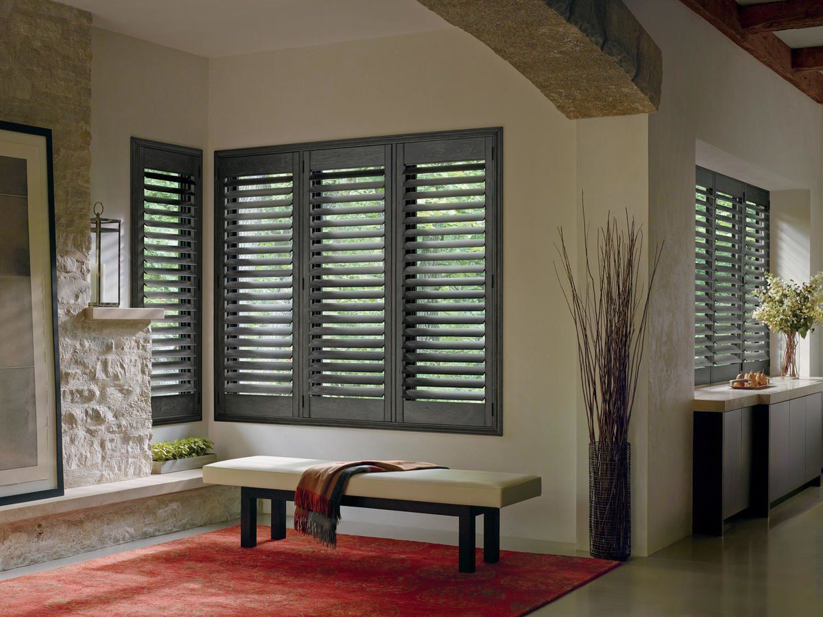 Dark-stained shutters on windows in a neutral-toned room. Red rug, bench, and plants add color.
