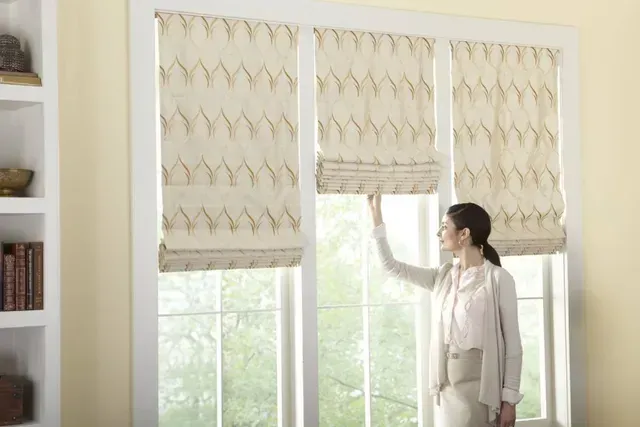 Woman adjusting a beige Roman shade in a window. Bright room with white trim and view of greenery.