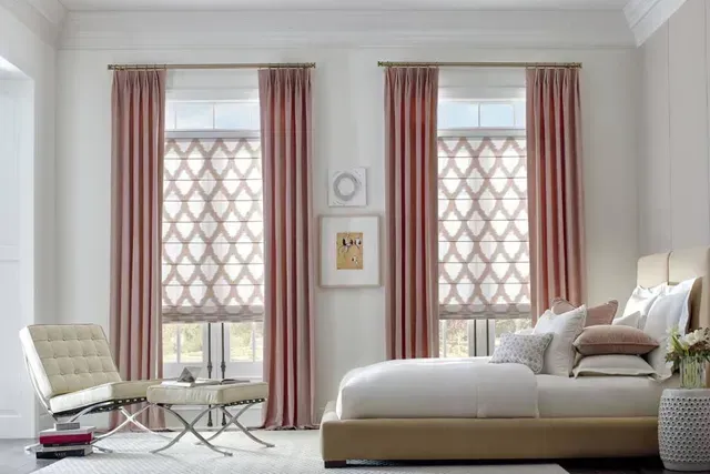 Bedroom with pink curtains, patterned shades, and a beige bed; a white chair sits nearby.
