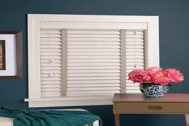 Window with white blinds and pink flowers on a table. The wall is dark blue.