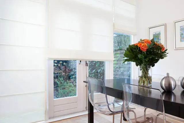 Dining area with white blinds, clear chairs, black table, flowers in vase.