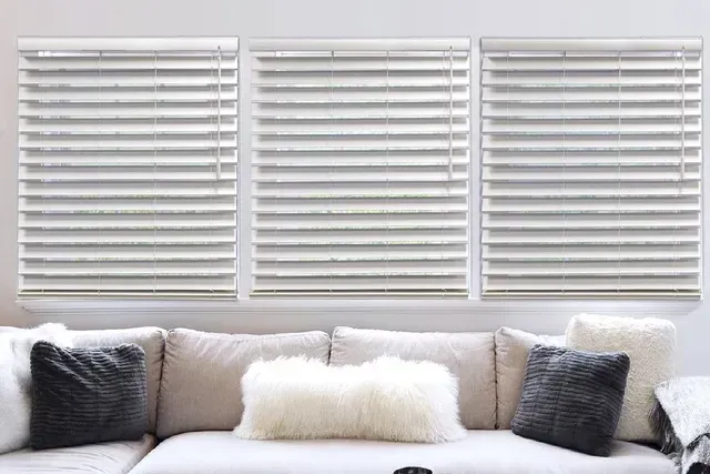 Three white-blinded windows above a cream-colored couch with decorative pillows.