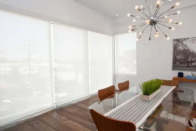 Dining room with large windows covered by white blinds, glass table, and modern chandelier.