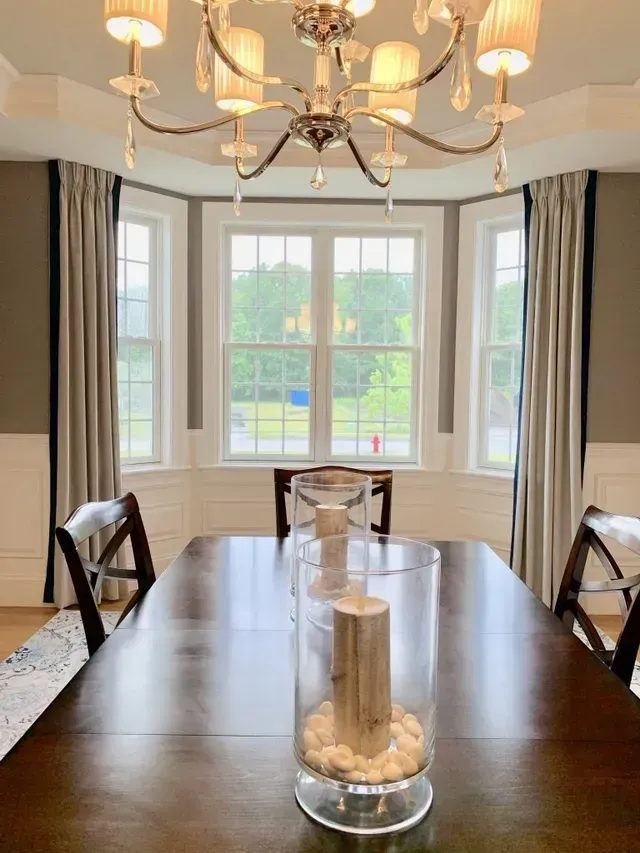 Dining room with dark wood table, chandelier, bay window, light curtains, and decorative candles.