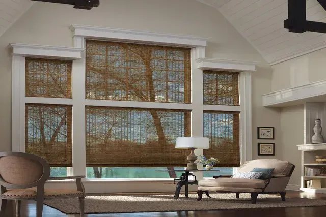 Living room with large windows featuring woven shades; furniture includes chair, chaise lounge, side table with lamp.