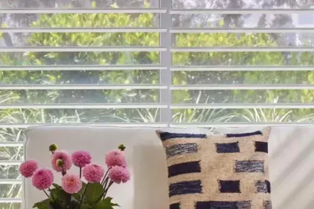White shutters partially open, pink flowers, and patterned pillow on a chair. Green foliage visible outside.