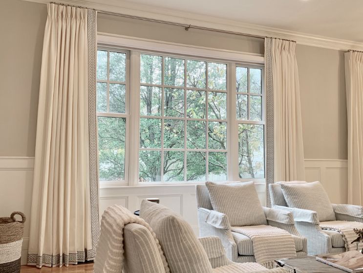 Living room window with white curtains, bordered in gray, framing a view of trees. White chairs and light-colored pillows.