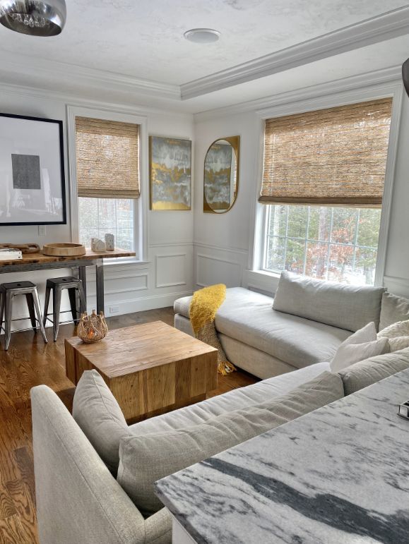 Living room with neutral tones: light gray couch, wooden coffee table, woven shades, and art on white walls.