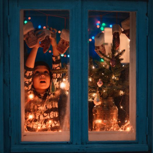 Child hanging mittens on string, lit by fairy lights inside a window with Christmas tree.