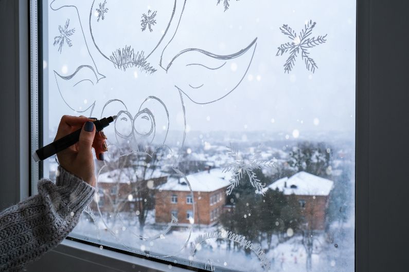 Person drawing a reindeer and snowflakes on a frosted window, overlooking a snow-covered town.