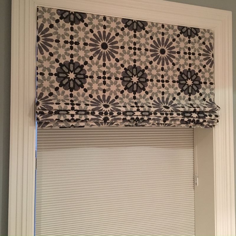 A patterned Roman shade partially covers a window with a white, pleated blind below.