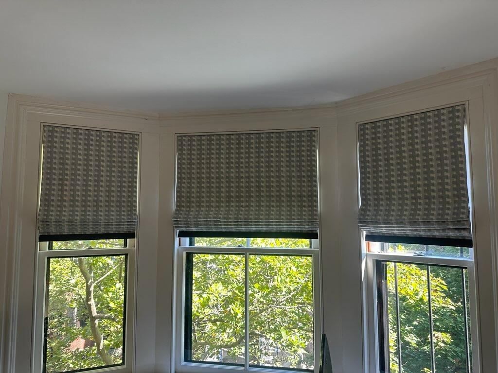 Three windows with gray patterned Roman shades, view of trees outside.