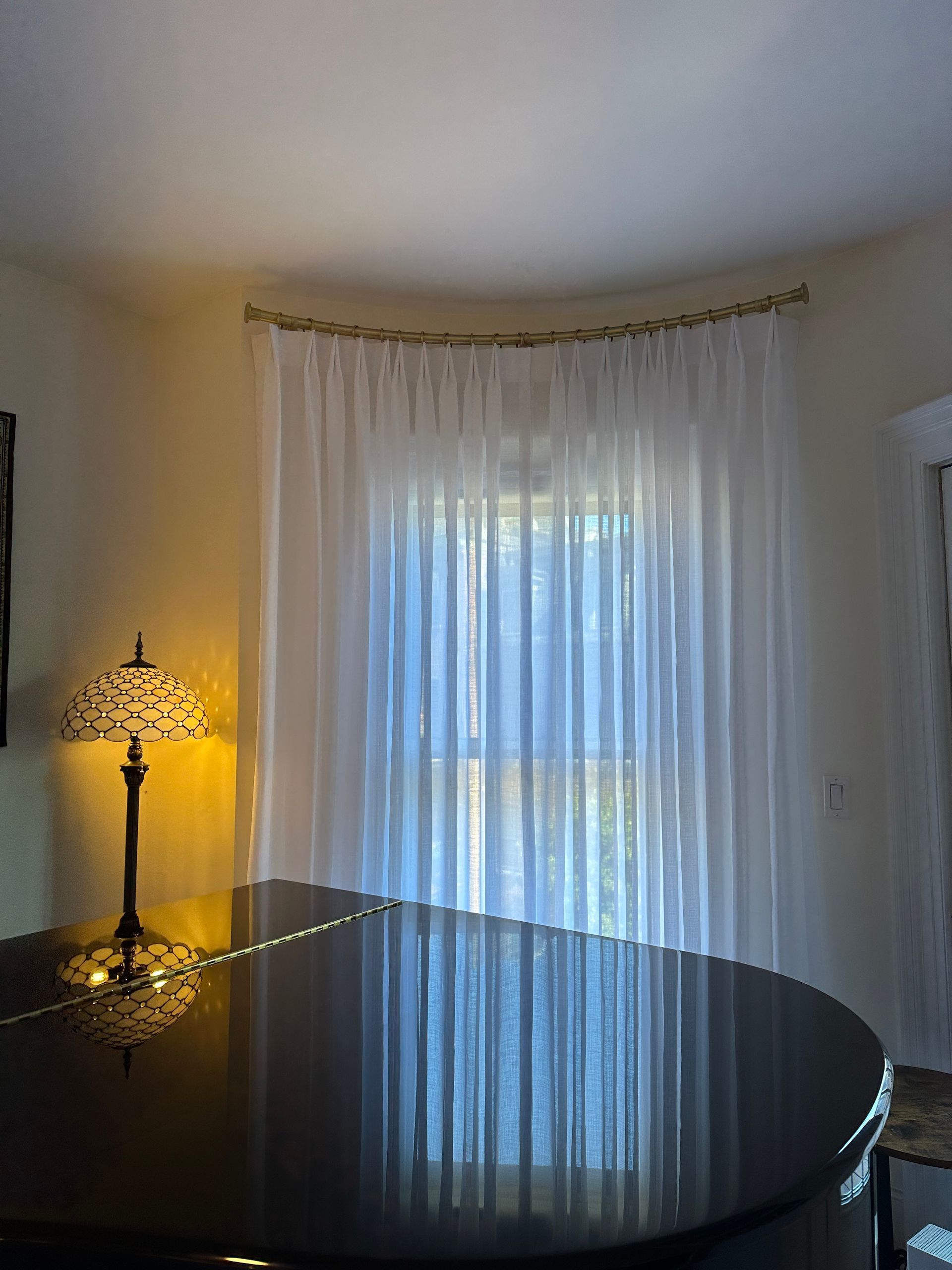 A piano with a lit lamp sits in front of a window with sheer white curtains, set in a light yellow room.