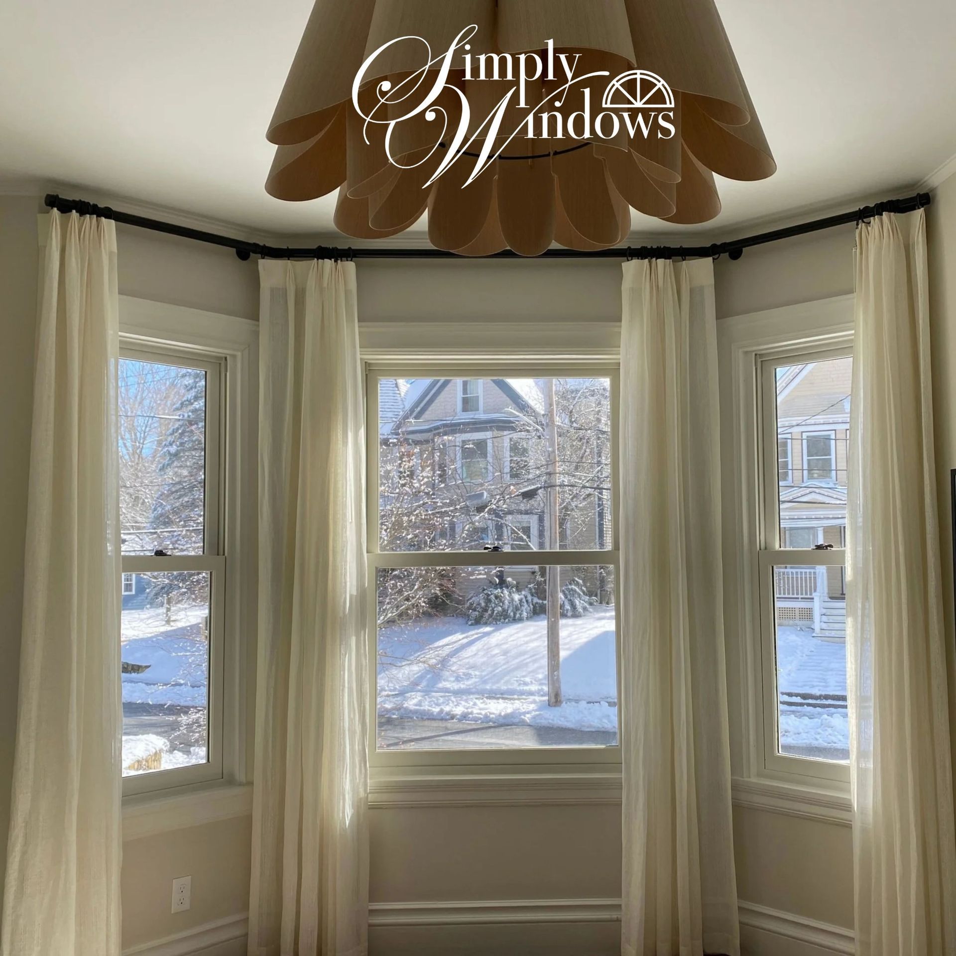 Bright bay window with white drapery overlooking a snowy suburban yard, with a “Simply Windows” logo above.