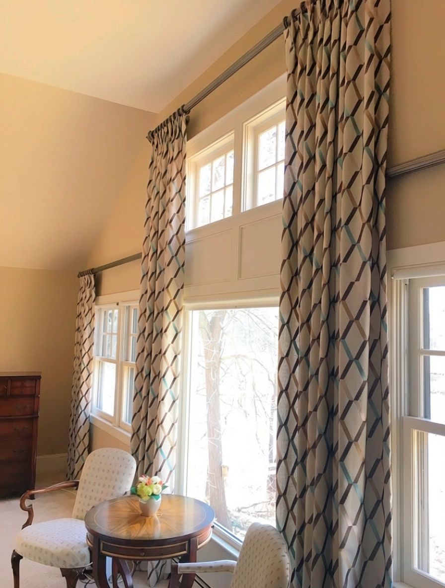 Tall windows with patterned curtains, a small table with chairs, and a chest of drawers.