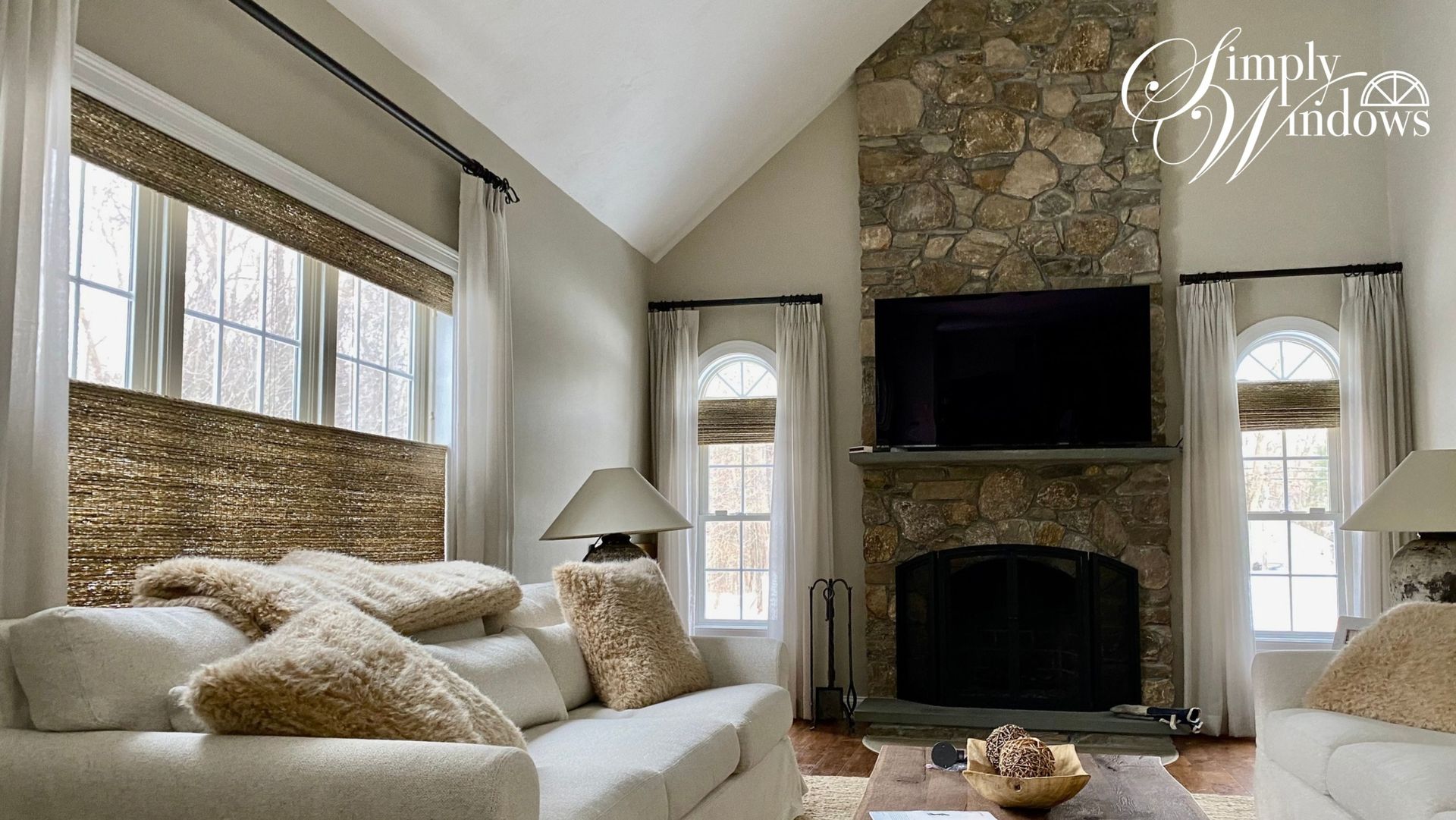 Cozy living room with stone fireplace, large TV, cream sofas, and tall windows with woven shades