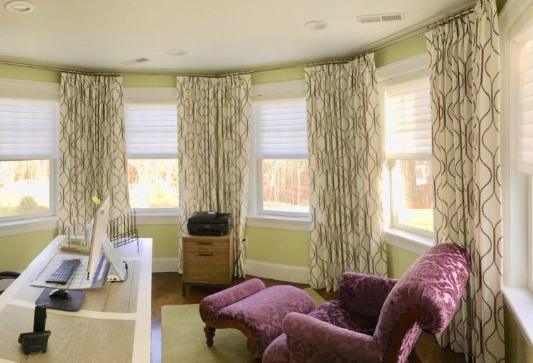 A bedroom with a bowed wall has drapes and honeycomb shades on the windows. 