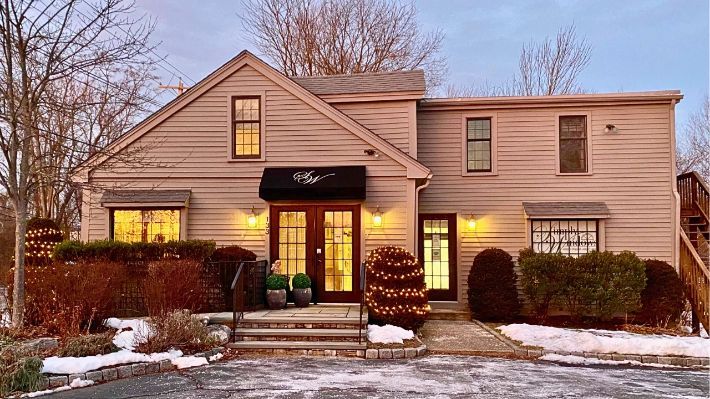 Beige building with a double-door entrance, small bushes, snow, and a Christmas tree; cloudy sky.