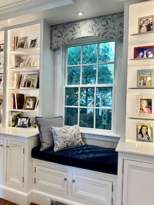 Built-in window seat with navy cushion and pillows, flanked by bookshelves and cabinets. Window with a leafy valance.