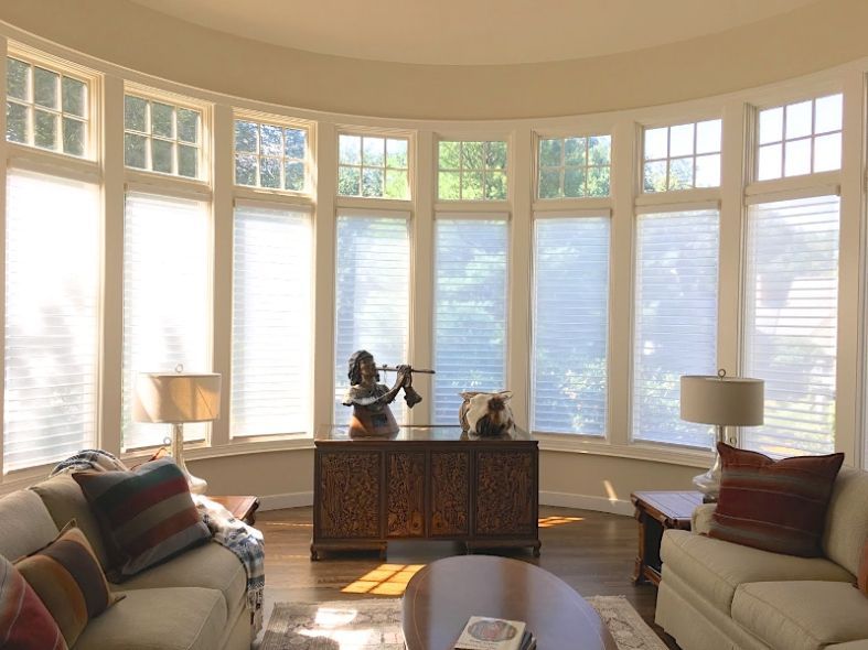Living room with curved windows, blinds, and a wood cabinet with statues. Two couches and end tables with lamps.