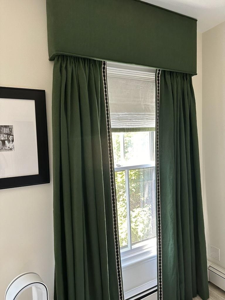 Green drapes frame a window with a Roman shade and trim, topped by a valance.